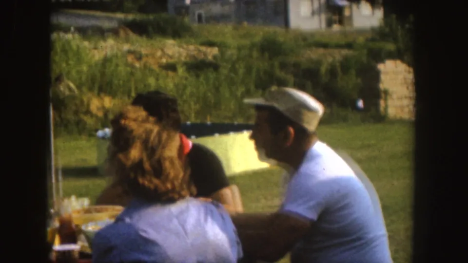 1955 Nebraska home-movie frame with adults seated at a picnic table in a sunny yard, including a man wearing a white cap and shirt.
