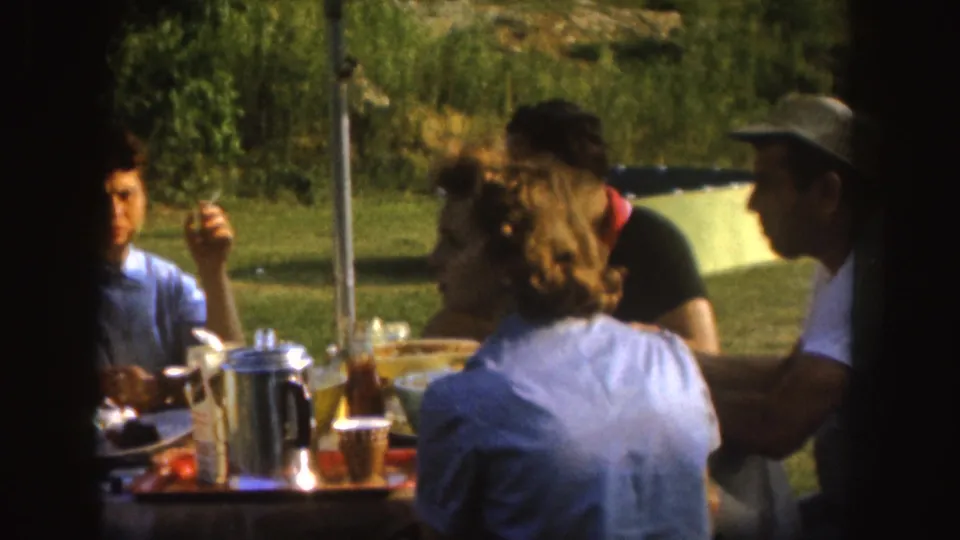 1955 Nebraska home-movie frame showing adults eating at a yard picnic table with plates, cups, tomatoes, and a metal coffee pot visible.