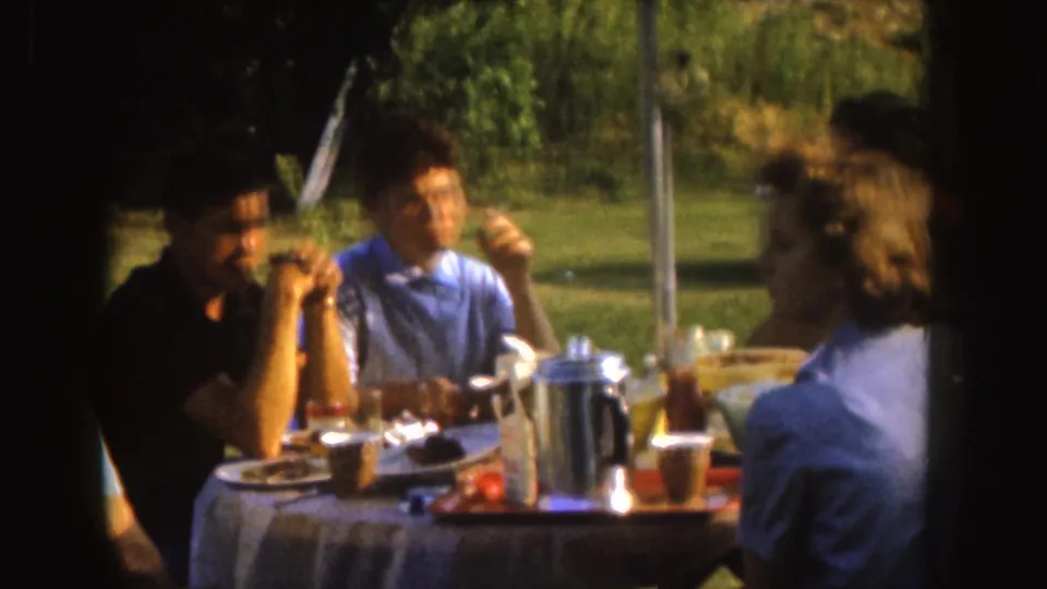 1955 Nebraska home-movie frame of adults at a picnic table with food, cups, and a metal coffee pot in a sunny residential yard.