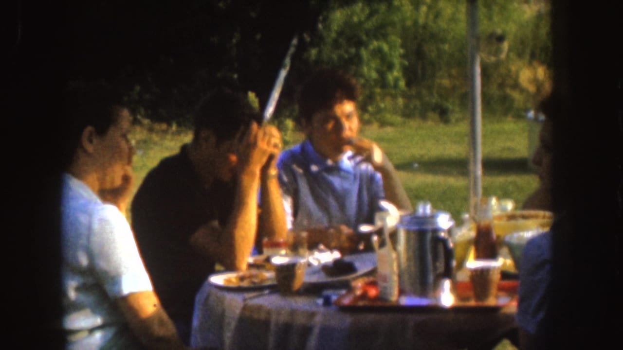 Adults seated around a picnic table with food, cups, and a metal coffee pot in a grassy park.