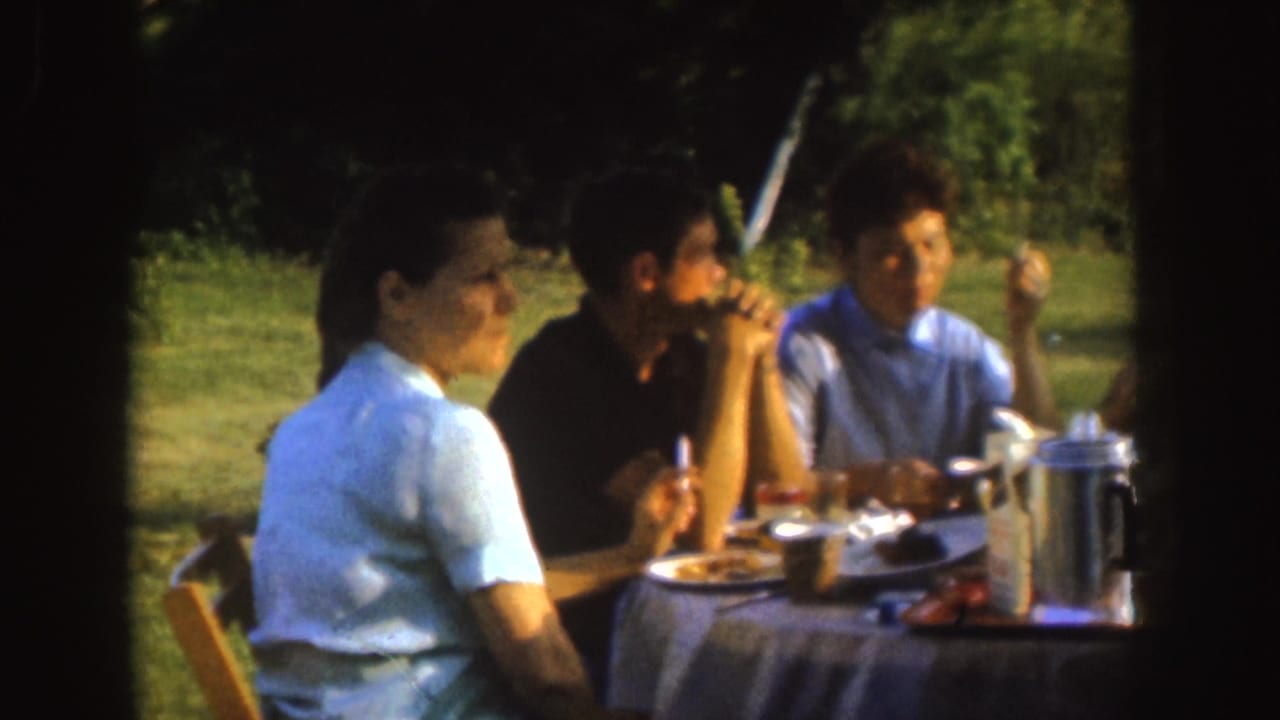 1955 Nebraska home-movie frame showing adults eating and talking at a small outdoor picnic table in a park setting.