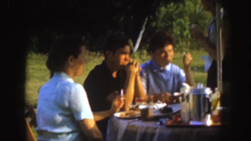 1955 Nebraska 8mm frame of adults sharing food and conversation at a picnic table, filmed in soft focus with trees behind them.