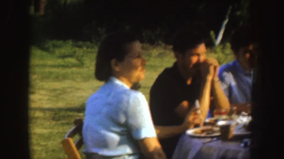 1955 Nebraska home-movie frame with adults seated at a park picnic table, one in a light shirt and another in a dark shirt near plates and cups.