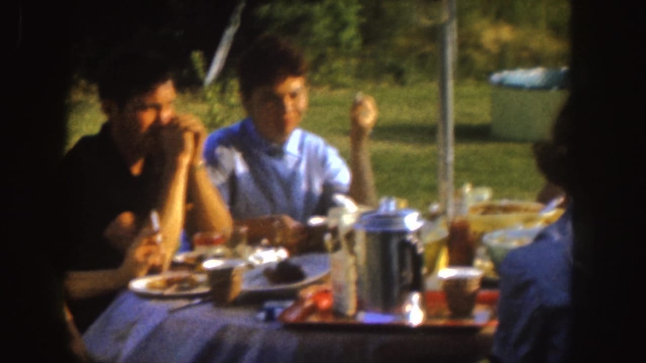 Adults at a 1955 Nebraska backyard table eat and converse, with a metal coffee pot, plates, cups, and lawn visible.
