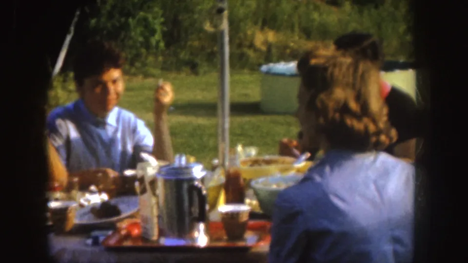 A woman in a light blue top sits across a backyard table during the 1955 Nebraska family meal, with dishes and a coffee pot nearby.