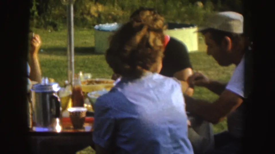 Adults gather around the backyard meal table in Nebraska, with a man in a light cap, a woman seen from behind, and tableware visible.