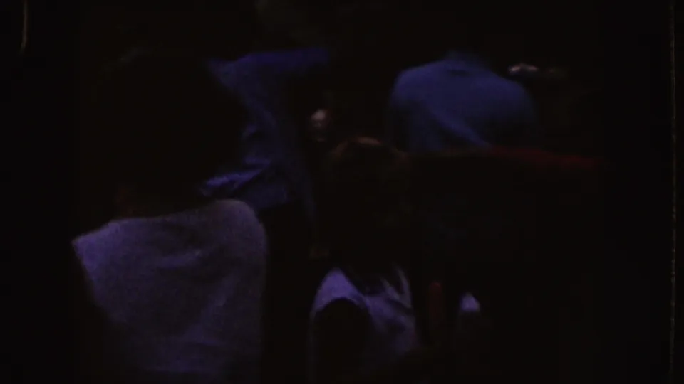 Nebraska 1955 home-movie frame showing a shadowed audience clustered in a dark indoor viewing space.