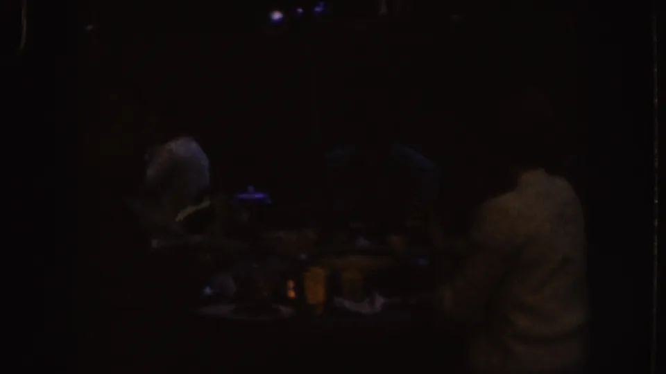 1955 Nebraska home-movie frame showing a dark indoor social scene with adults seated near drinks and glassware.