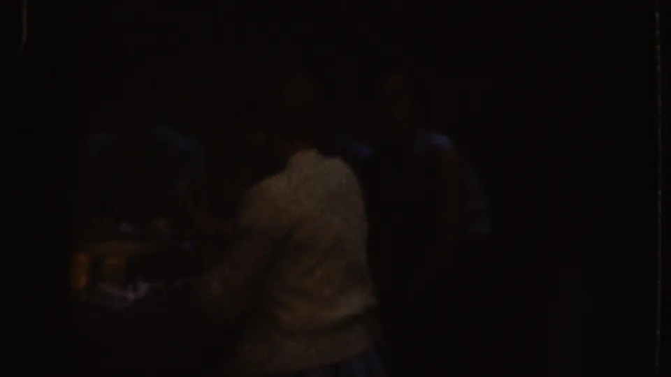 Low-light 1955 Nebraska 8mm frame showing adults gathered near a table, with one light-shirted figure most visible in shadow.