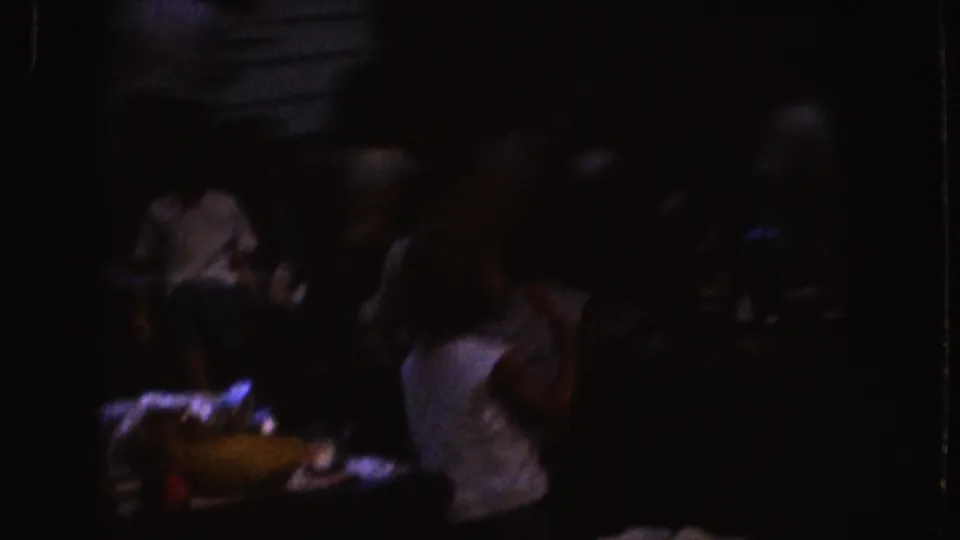 Low-exposure 1955 Nebraska frame showing seated figures at a table with food or dishes, filmed in grainy 8mm home-movie style.