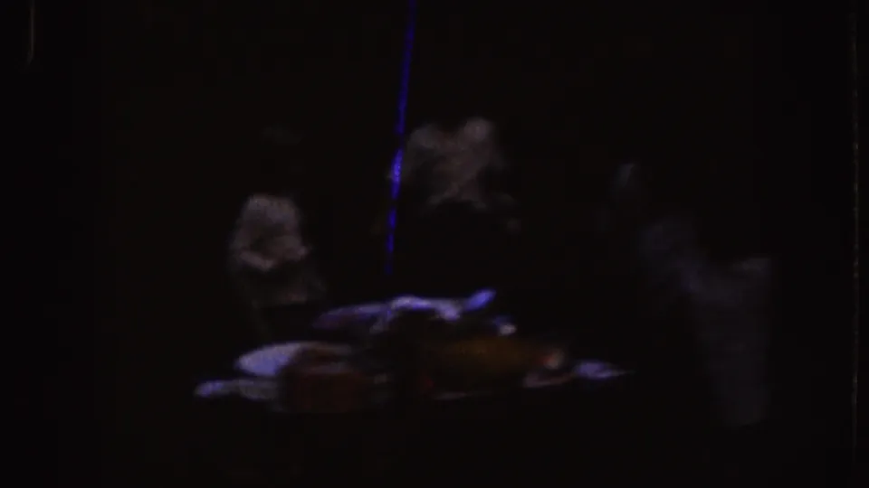 1955 Nebraska home-movie frame showing a dim table with food or serving items and nearby adults in deep shadow.