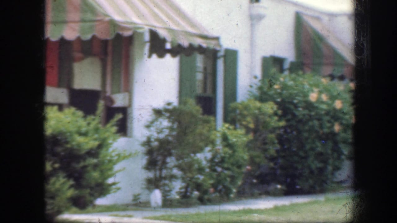 White house exterior with striped awning, green shutters, bushes, lawn, and dark window framing.