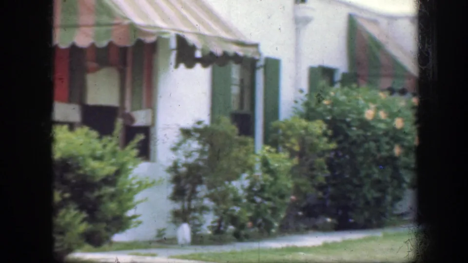 1945 Florida 8mm frame showing a white-sided house with striped awning, green shutters, shrubs, and a small lawn seen through dark framing.