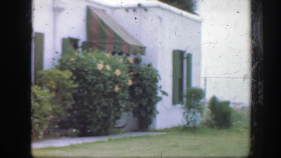 1945 Florida home-movie frame of a white house exterior with green shutters, striped awning, bushes, lawn, and soft film grain.