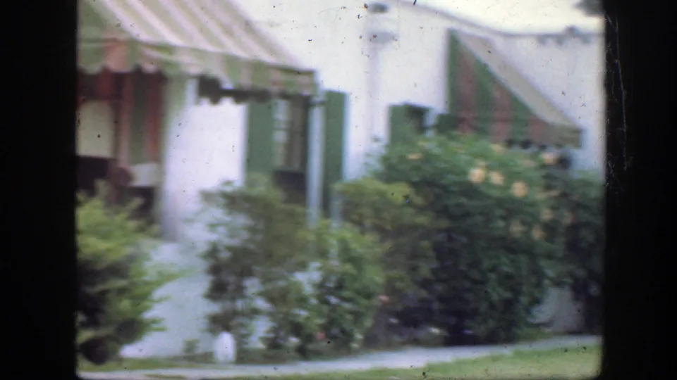 1945 Florida 8mm frame with a white-sided house, striped awning, green shutters, shrubs, lawn, walkway, and dark vertical frame edge.