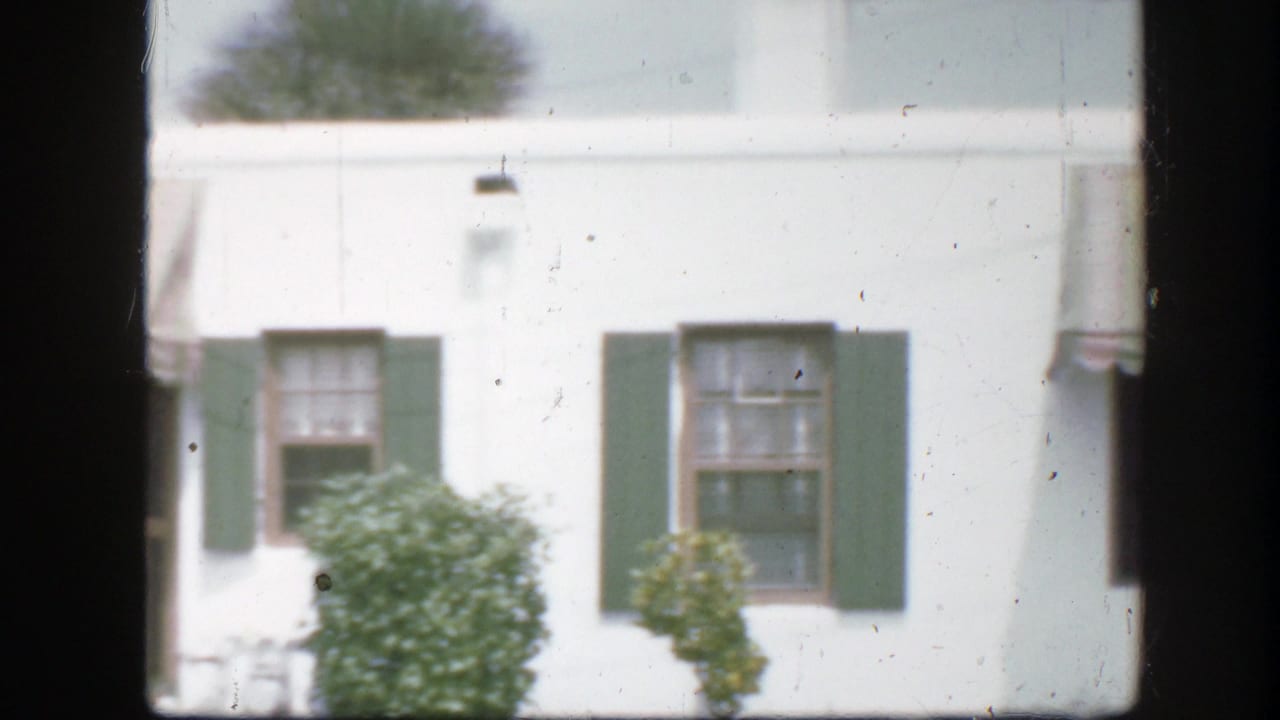 White house exterior with green shutters, shrubs, and a partial striped awning.