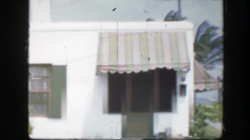 1945 Florida frame with a white house exterior, green door, striped awning, dark window opening, and palm fronds nearby.