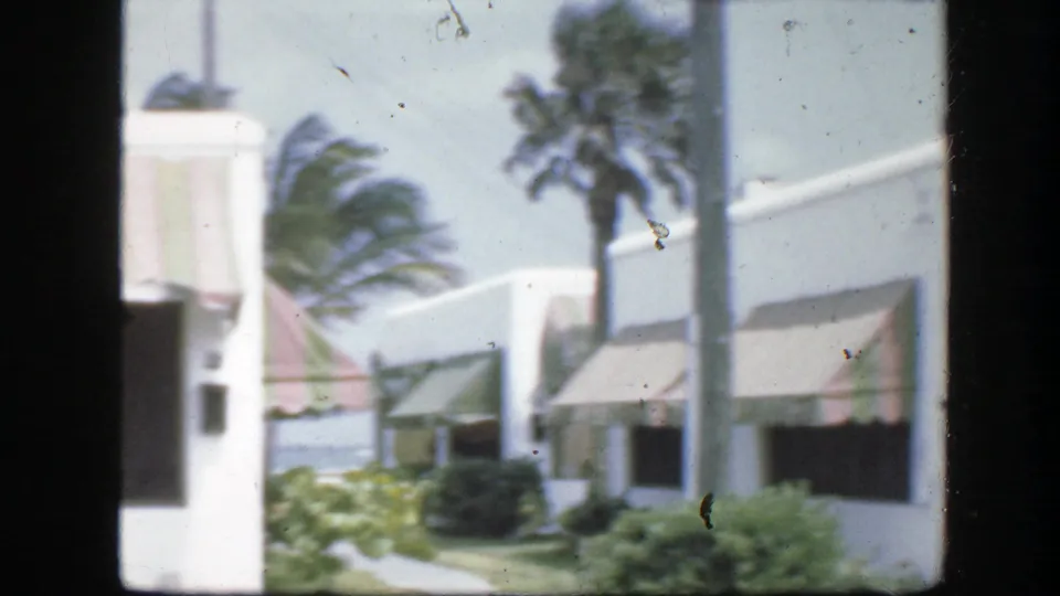 1945 Florida home-movie view of white residential buildings with awnings, palm trees bending in wind, shrubs, and a walkway.