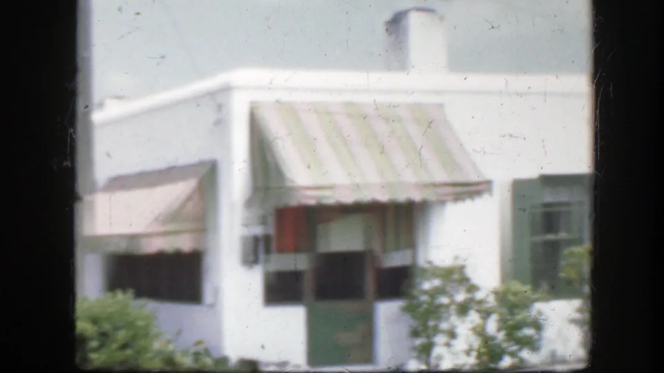 1945 Florida frame showing a white house corner with chimney, striped awning, green door, window, shrubs, and soft 8mm grain.