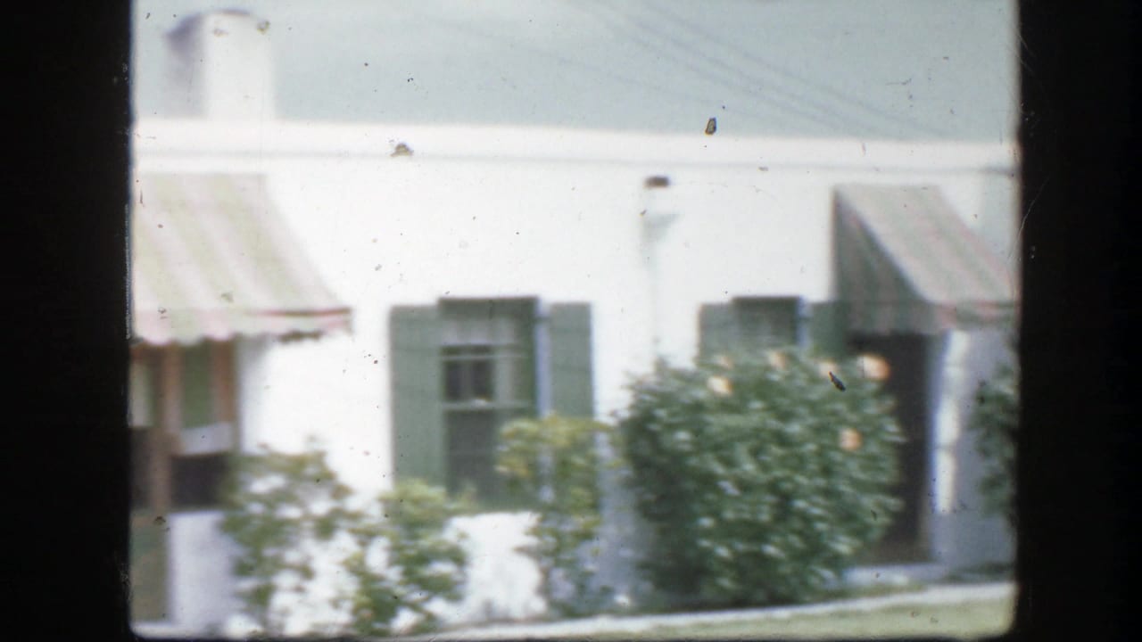 White house with dark shutters, striped awning, shrubs, and lawn seen from a moving camera.