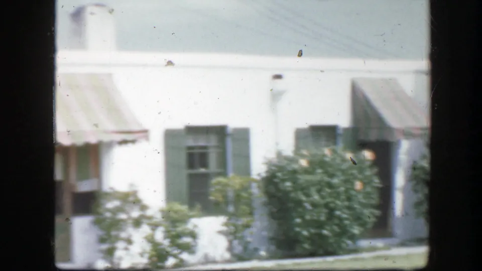 1945 Florida 8mm frame showing a white residential house with green shutters, striped awning, shrubs, and a sunlit lawn.
