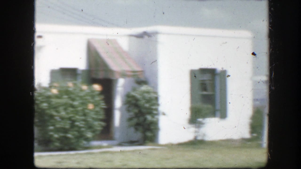 White house facade with striped awnings, dark shutters, large shrubs, and grass in front.