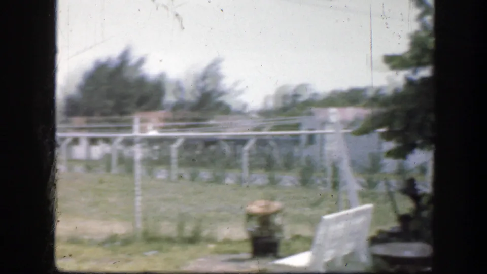 1945 Florida home-movie frame showing a fenced grassy field, low rails, trees, shrubs, and a bright foreground object near the roadside.