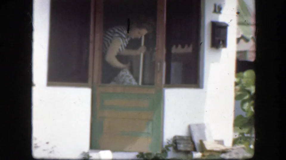 The woman remains partly inside the Florida house doorway, with the green door, wall mailbox, and sunlit plants visible around the entrance.