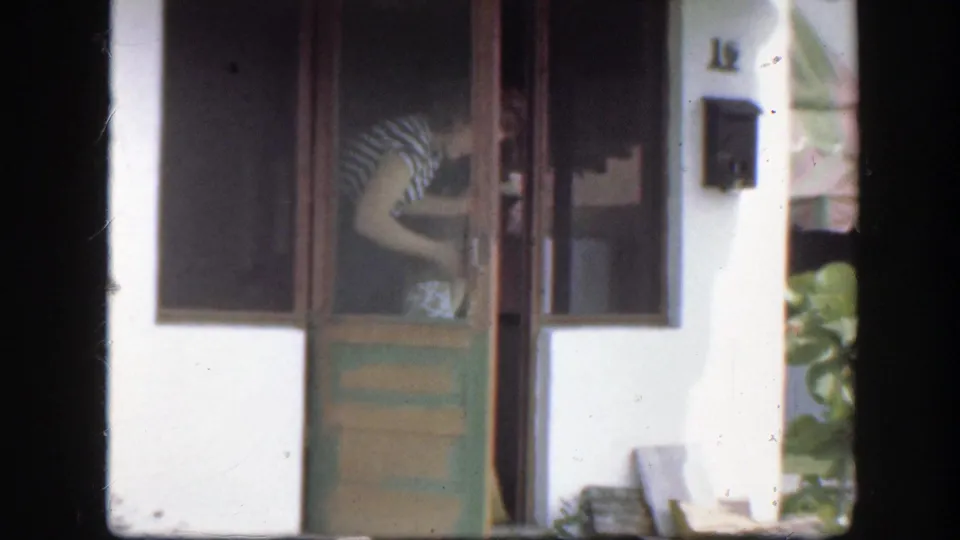 A woman in a striped dress bends near the green doorway with a broom, seen through soft, grainy 8mm home-movie color.
