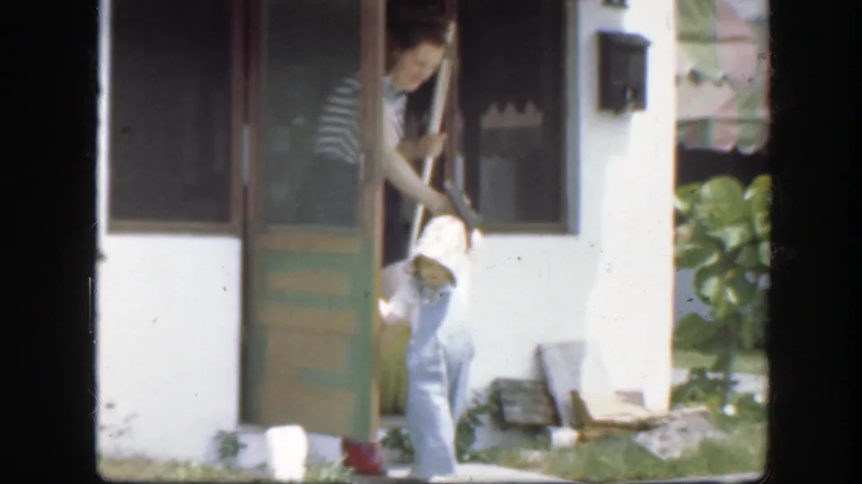 At the white house entrance, the woman reaches toward a small child near the green door, with plants and a wall mailbox in view.