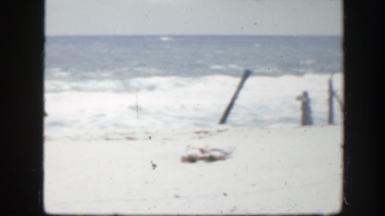 1945 Florida home-movie frame showing pale beach, rolling surf, and weathered wooden posts near the shoreline.