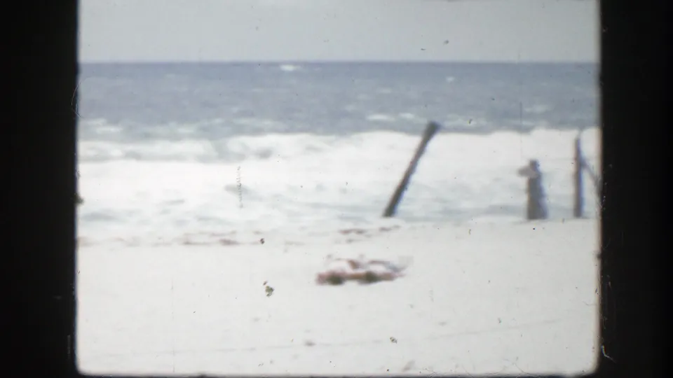1945 Florida home-movie frame showing pale beach, rolling surf, and weathered wooden posts near the shoreline.
