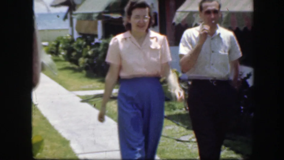 In 1940s Florida footage, an adult couple walks toward the camera along a resort-area sidewalk.