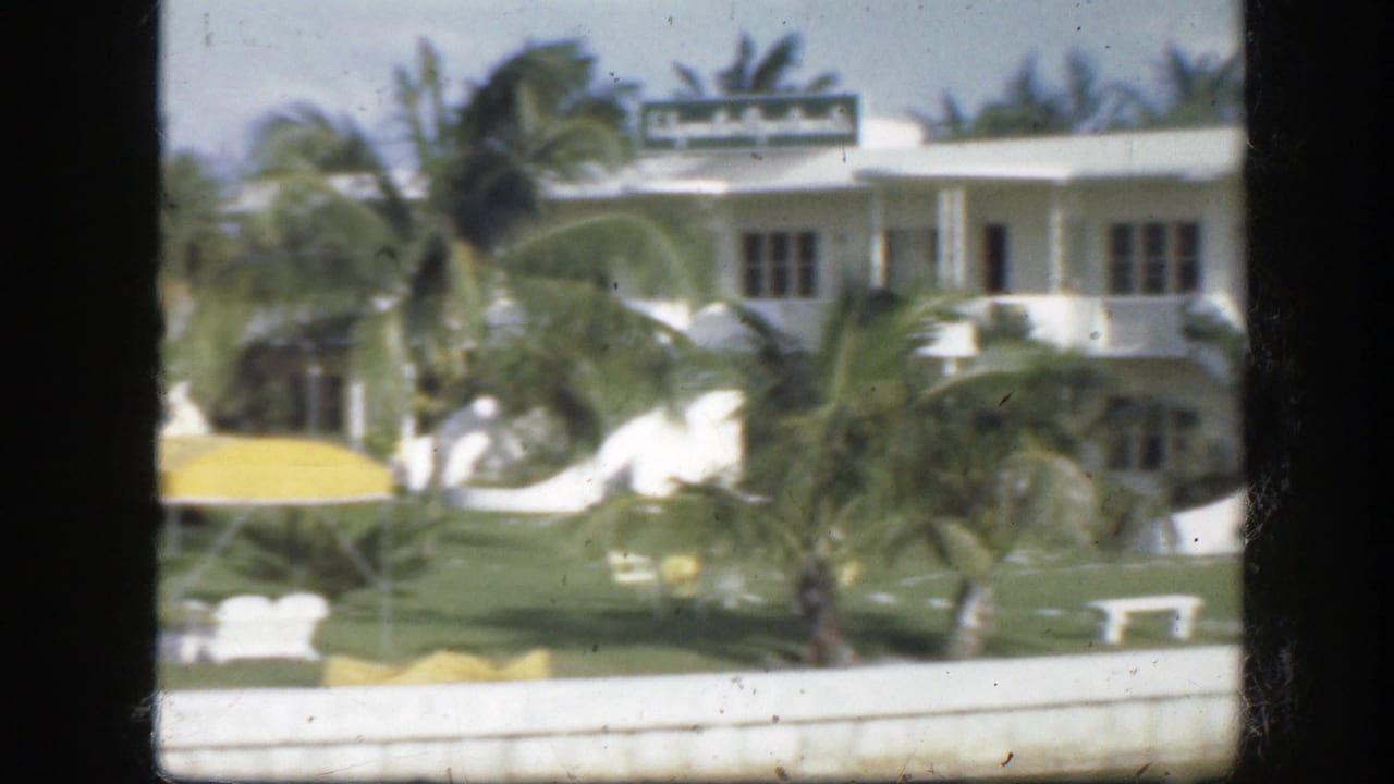 1945 Florida home-movie view of a white waterfront building with palms, lawn furniture, and a yellow awning.