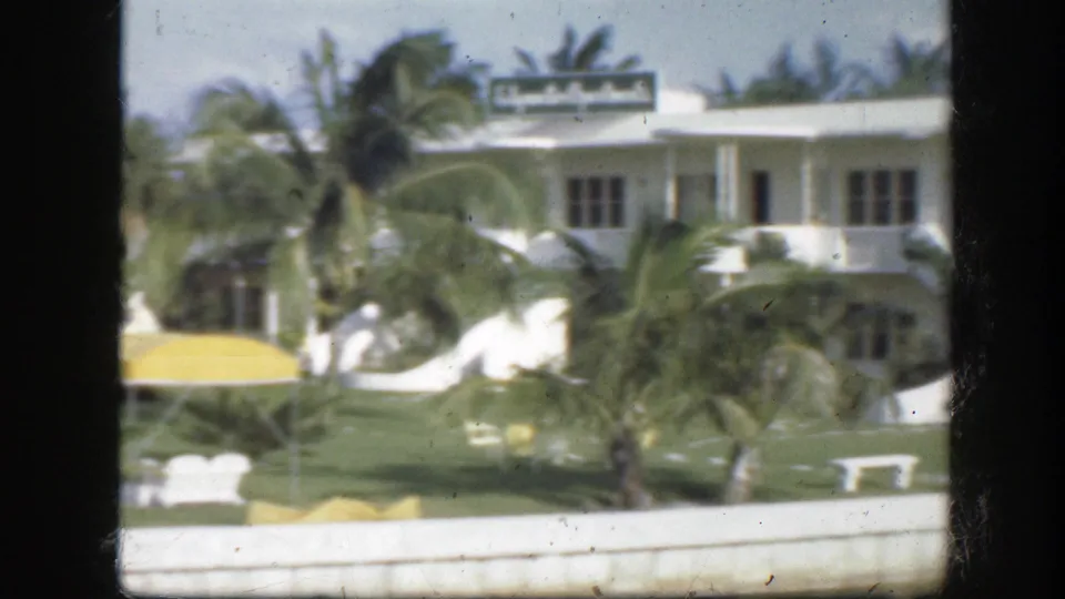 1945 Florida home-movie view of a white waterfront building with palms, lawn furniture, and a yellow awning.