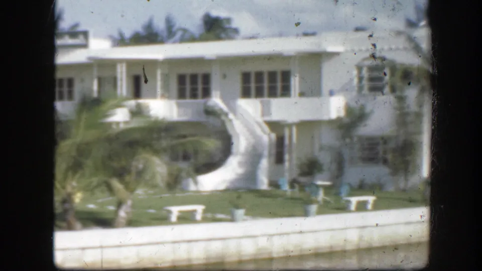1945 Florida view of a white waterfront building with a curved staircase, palms, and manicured lawn.