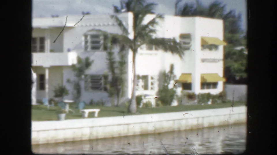 1945 Florida home-movie frame showing a rounded white building with yellow awnings beside a waterfront lawn.
