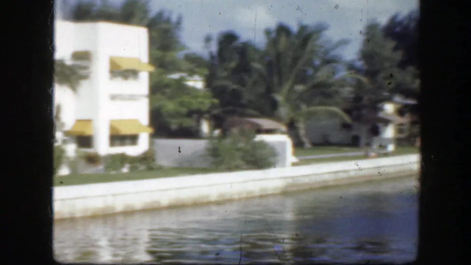 1945 Florida waterfront view with white buildings, yellow awnings, palm trees, and calm water in the foreground.