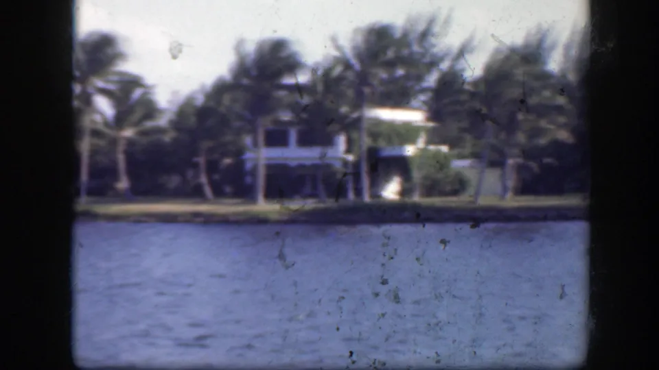 1945 Florida shoreline frame showing a pale waterfront house among palms, viewed from a boat across the channel.