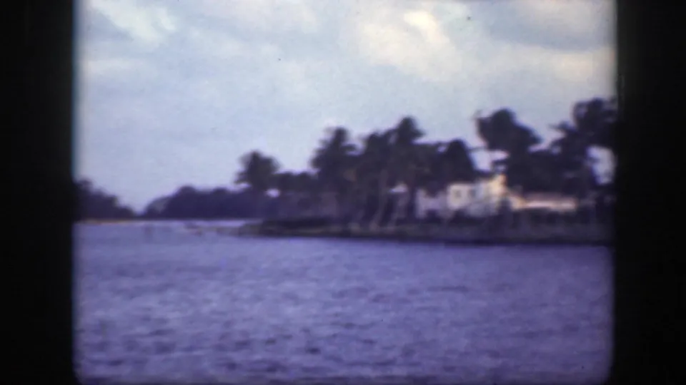 1945 Florida frame looking down a waterway with palm trees and houses along the right shoreline.