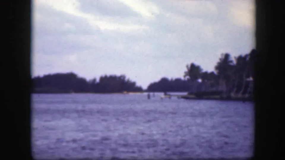 1945 Florida 8mm frame showing calm blue water, a low palm-lined shoreline, distant trees, and a soft cloudy sky.