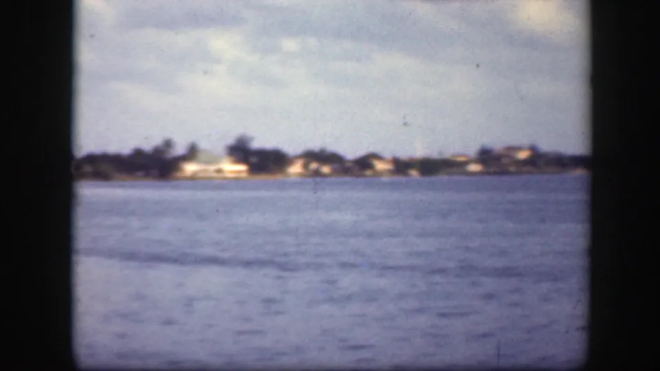 1945 Florida 8mm frame showing a calm waterway with distant shoreline houses, trees, and muted blue sky.