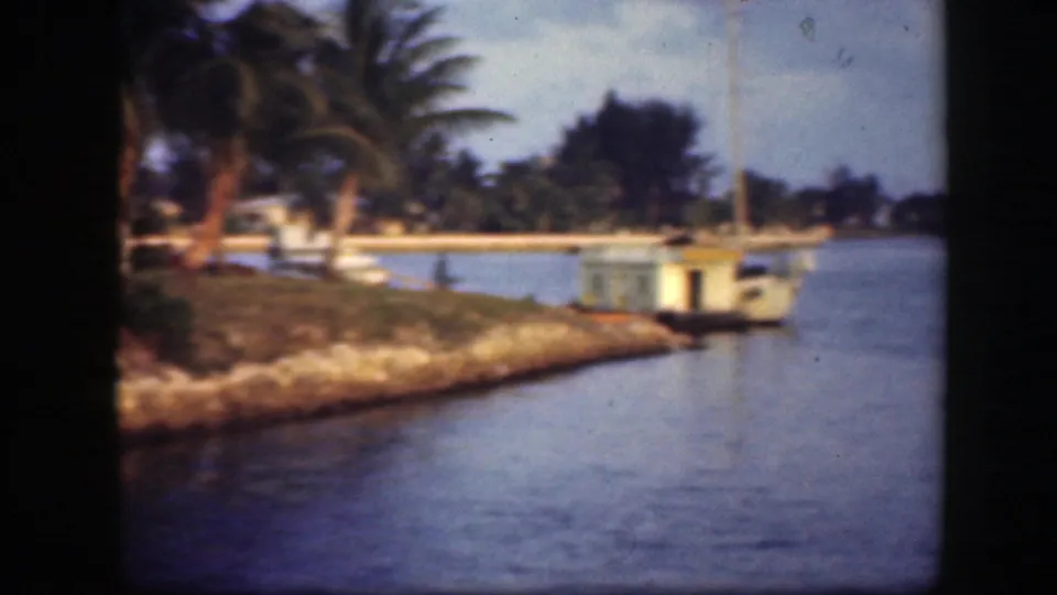 1945 Florida 8mm frame showing palms on a narrow waterside bank and a small dock or boathouse by calm water.
