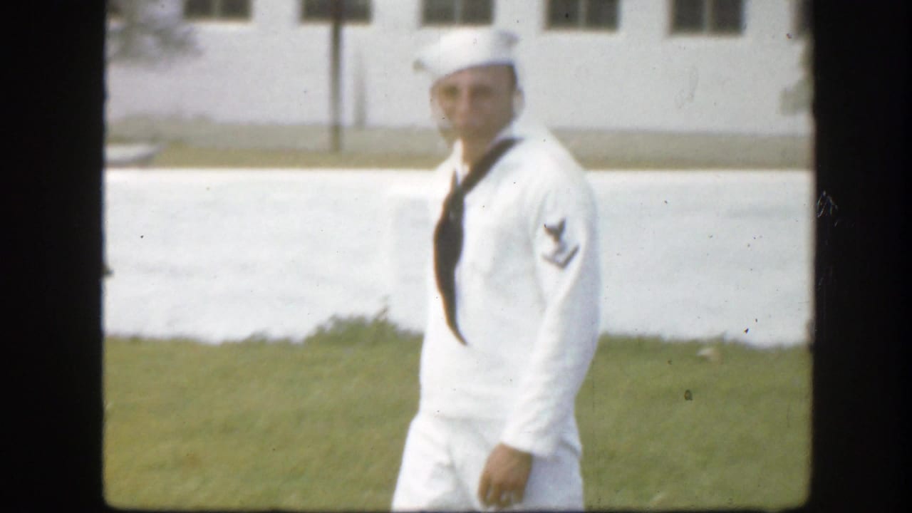 Sailor in a white uniform walking on grass beside a pale building.