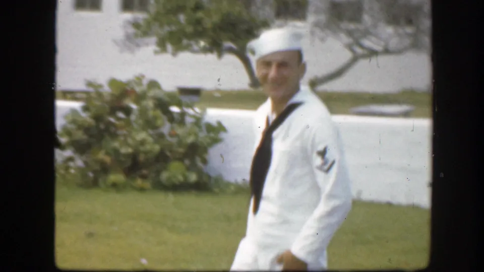 Smiling adult sailor in white uniform walks outdoors with shrubs and a pale wall behind him in Florida, 1945.