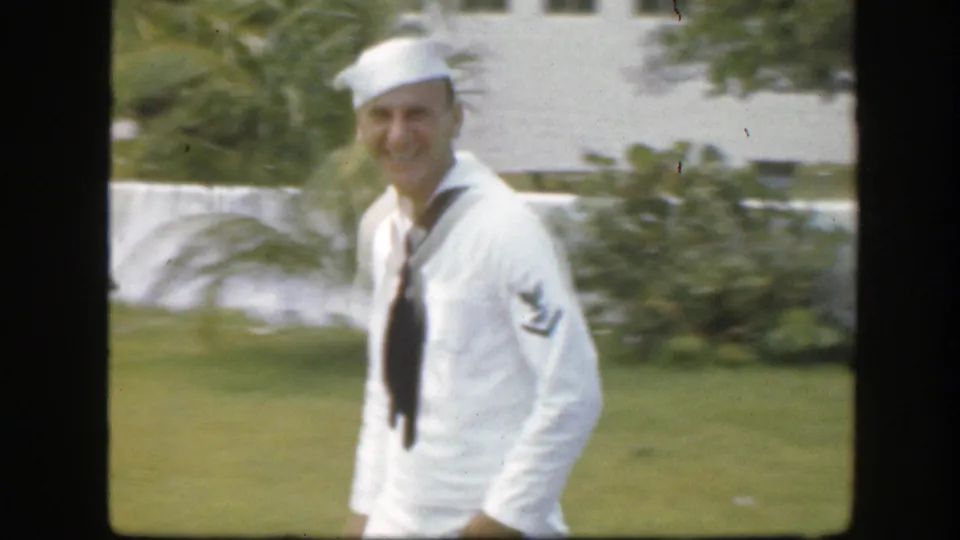 Adult sailor smiles while walking near the camera, with palm foliage and lawn visible in the 1945 Florida footage.