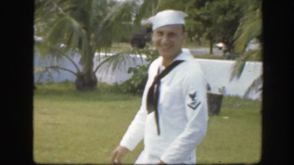 Smiling sailor in a white uniform walks past palm trees and grass in the supplied 1945 Florida home-movie clip.