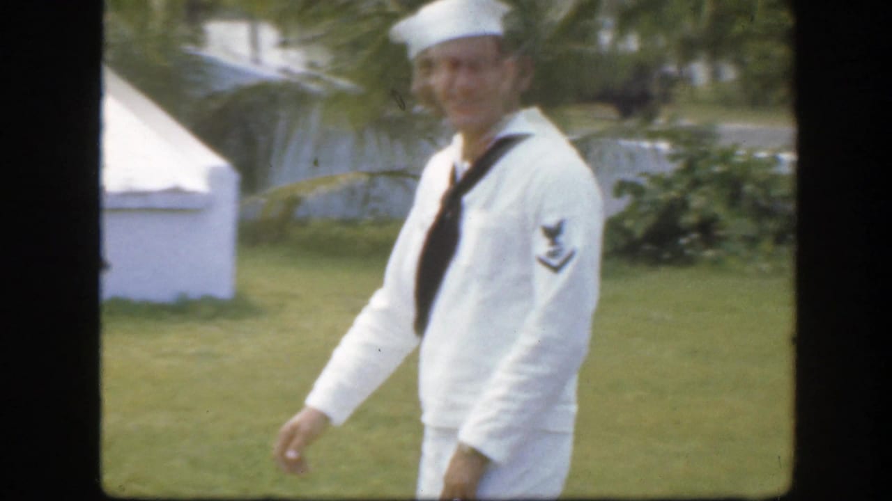 In supplied 1945 Florida metadata, a sailor in a white uniform walks on a sunlit lawn near white buildings and greenery.