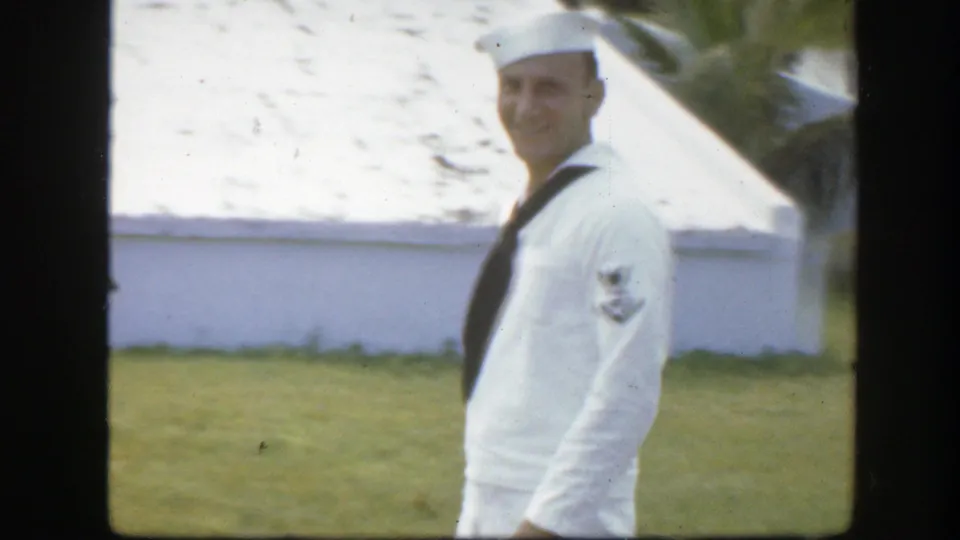 A smiling sailor in a white uniform stands on grass beside a white building, consistent with the supplied Florida home-movie setting.