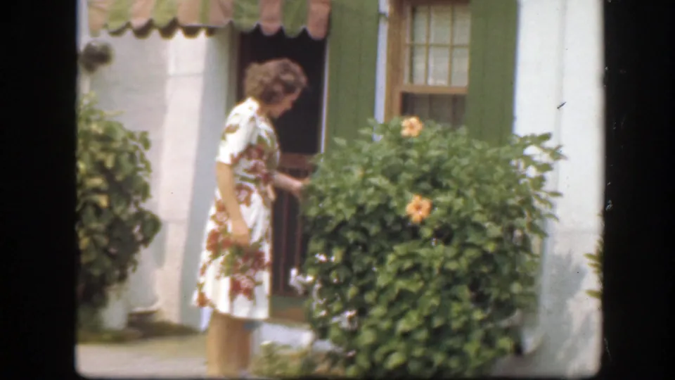 A woman in a floral dress stands by a doorway and leafy flowering bush outside a white house with green shutters.